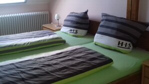 A room with two single beds, each with a green mattress and striped pillows. There is a small bedside table with a lamp, and a radiator is visible in the corner.