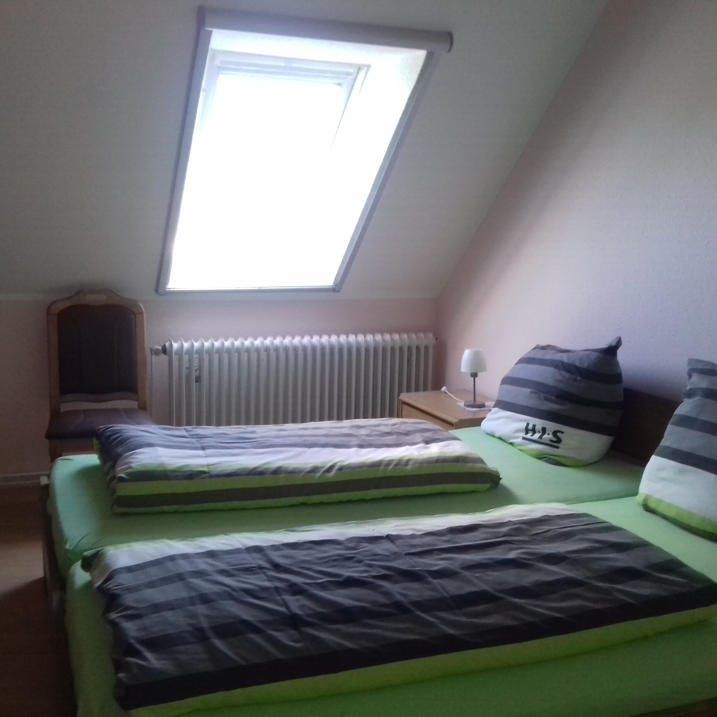 A bedroom with two beds, a skylight, a radiator, a nightstand with a lamp, and striped pillows.