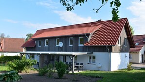 A two-story house with a red-tiled roof, a covered patio with outdoor furniture, and a garden area with various plants.