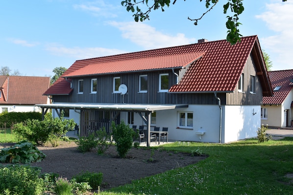 A two-story house with a red-tiled roof, a covered patio with outdoor furniture, and a garden area with various plants.
