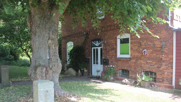 A brick building with a white door, a small porch, and a tree in the foreground. - Charming family and pet friendly apartment in rural idyll - pure relaxation (Kiel Pries)
