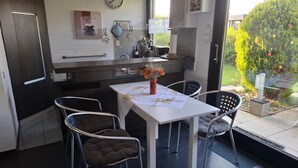 Una cocina moderna con zona de comedor, que presenta una mesa blanca con mantel floral, dos sillas acolchadas y un florero con flores secas.