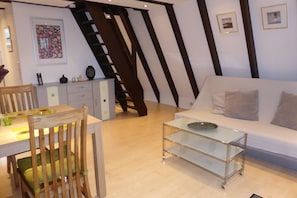 Una sala de estar moderna con una mesa de centro de vidrio, un sofá blanco, una zona de comedor con una silla de madera y una escalera con barandillas de madera oscura.
