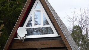 Una cabaña pequeña de forma triangular, con fachada blanca, antena parabólica y terraza de madera.