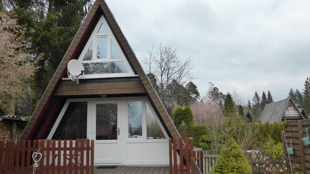 Una cabaña de madera triangular con puerta y ventanas blancas, rodeada por una terraza de madera y una valla de piquetes.