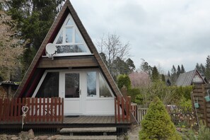 Una cabaña de madera triangular con puerta y ventanas blancas, rodeada por una terraza de madera y una valla de piquetes.