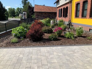 Un chemin pavé mène à un jardin où l’on trouve différentes espèces de plantes, ainsi qu’à un bâtiment en pierre surmonté d’un toit recouvert de tuiles rouges.