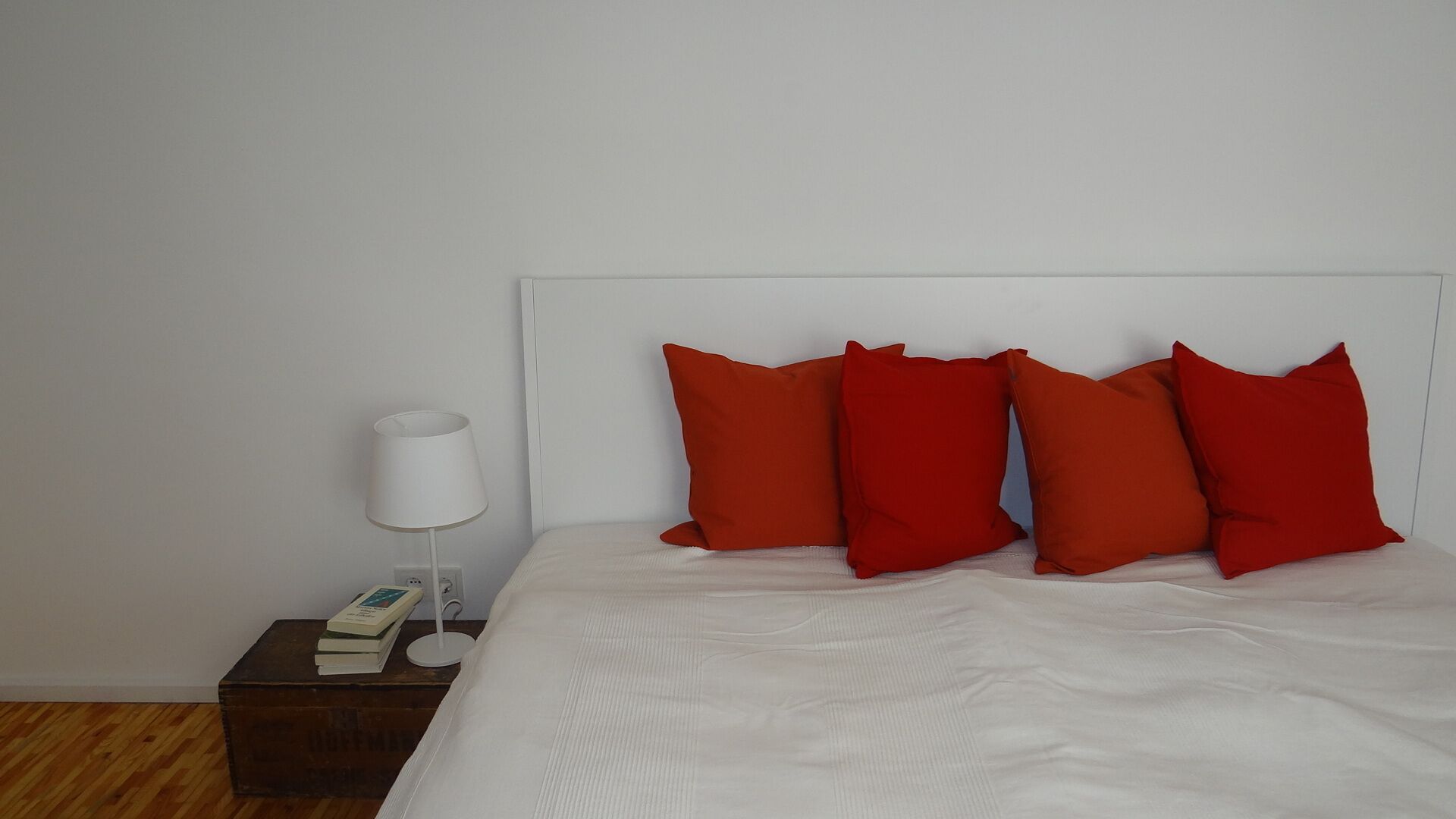 A neatly made bed with four red pillows and a white headboard. A white bedside lamp and a small wooden nightstand with a book are visible.