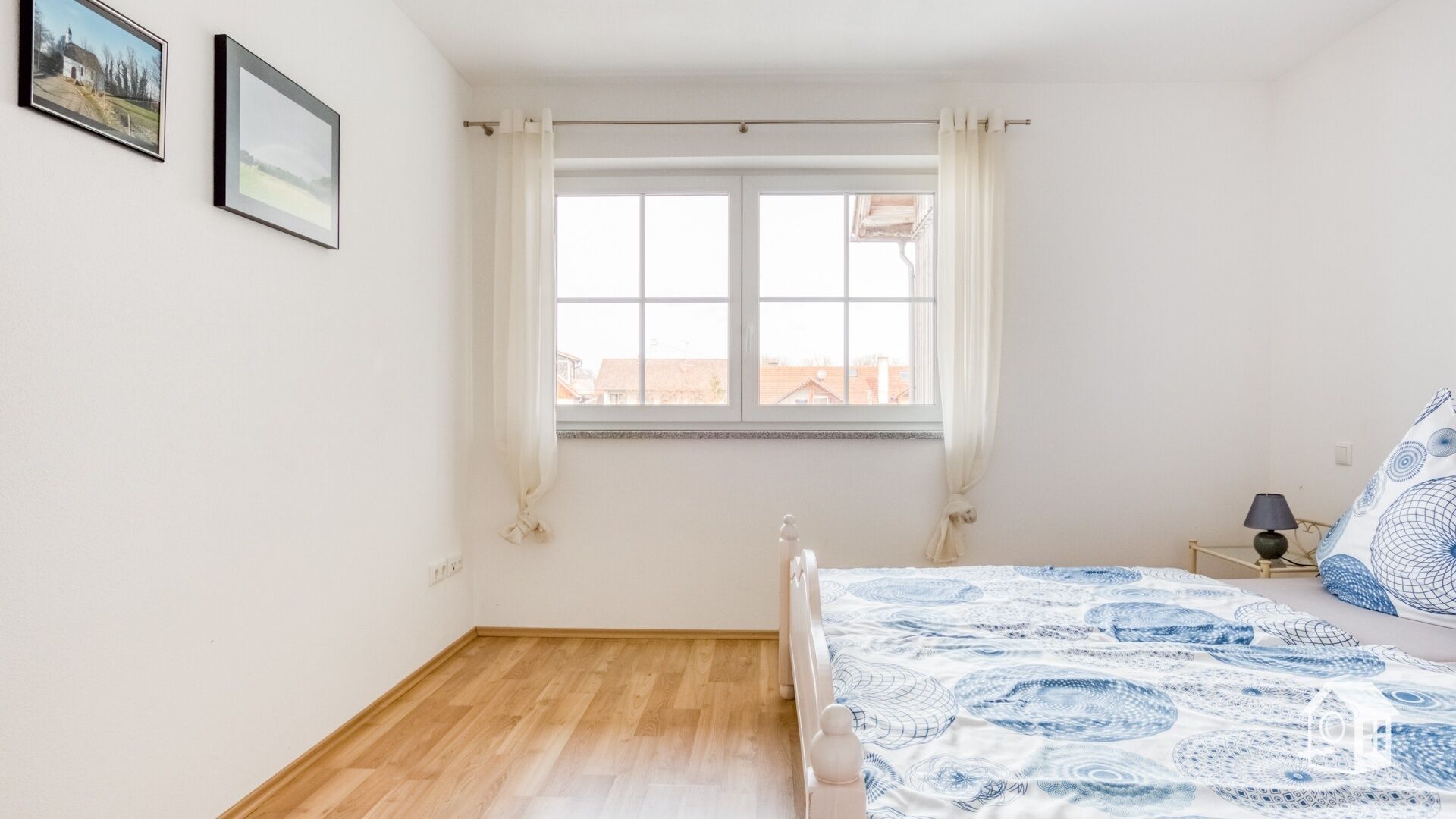 A bedroom with a wooden floor, a bed with a blue and white patterned bedspread, a bedside table with a lamp, and two framed pictures on the wall.