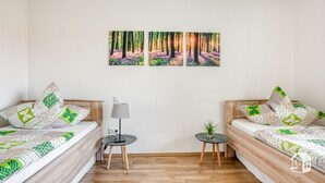 A bedroom with two beds, a bedside table with a lamp, a small table with a plant, and three wall-mounted paintings of a forest scene.