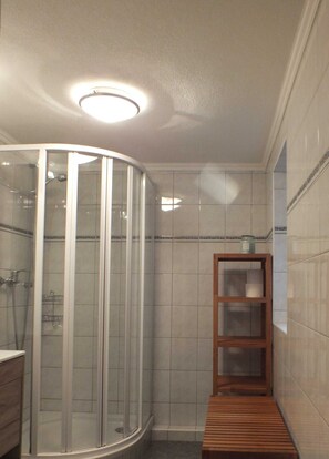 A bathroom with a glass shower enclosure, a wooden bench, and a shelf with toiletries.