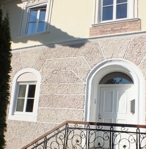 Une façade d’immeuble avec un mur en pierre à texture, une porte blanche et deux fenêtres à encadrements blancs.