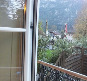 Une vue depuis un balcon doté d’une balustrade en bois, donnant sur un village aux bâtiments traditionnels, avec une chaîne de montagnes en arrière-plan.