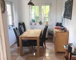 A dining area with a wooden table and chairs, a window with a view of greenery, and a bookshelf with books and a lamp.