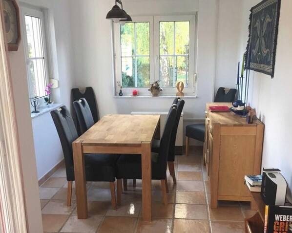 Un coin repas avec une table et des chaises en bois, une fenêtre donnant sur un paysage verdoyant, et une bibliothèque contenant des livres et une lampe.