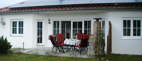 A white house with a tiled roof, a stone chimney, and a patio with a table and chairs.