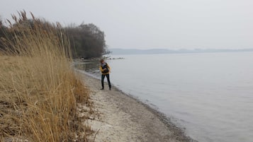 A person standing by a calm body of water with dry grass and trees in the background.