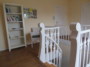 A staircase with white balusters, a wicker chair, and a bookshelf with books. - Apartment Mönchgut - Pension Waldrose (Sellin)