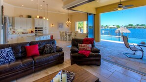 A spacious living area with a large window overlooking a pool and water view. There are two leather sofas with patterned cushions and a coffee table in the center. - Luxury villa in a prime location with an oversized pool and whirlpool (Cape Coral)