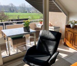 A balcony with a black leather chair, a table with a potted plant, wicker chairs, and a view of a green field.