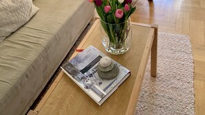 Un salon comprenant un canapé, un fauteuil, une table basse avec un magazine, un vase de fleurs et une grande fenêtre avec des rideaux.