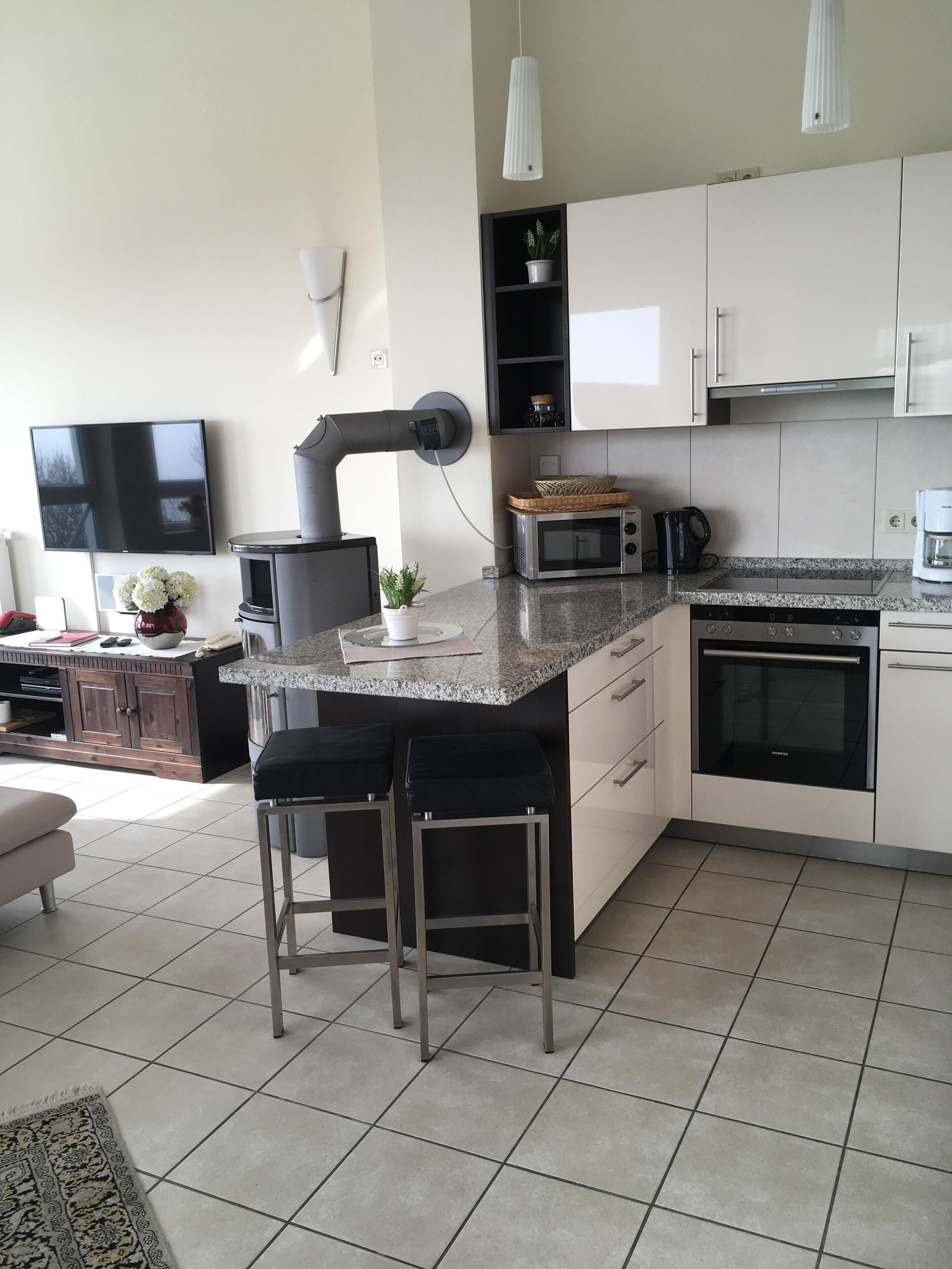 A modern kitchen with white cabinets, a granite countertop, a built-in oven, a microwave, and a coffee maker. There are two bar stools and a small island with a sink.