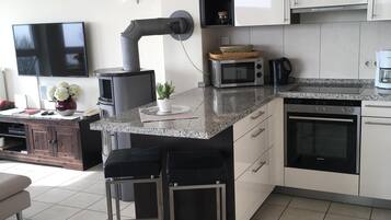 A modern kitchen with white cabinets, a granite countertop, a built-in oven, a microwave, and a coffee maker. There are two bar stools and a small island with a sink.