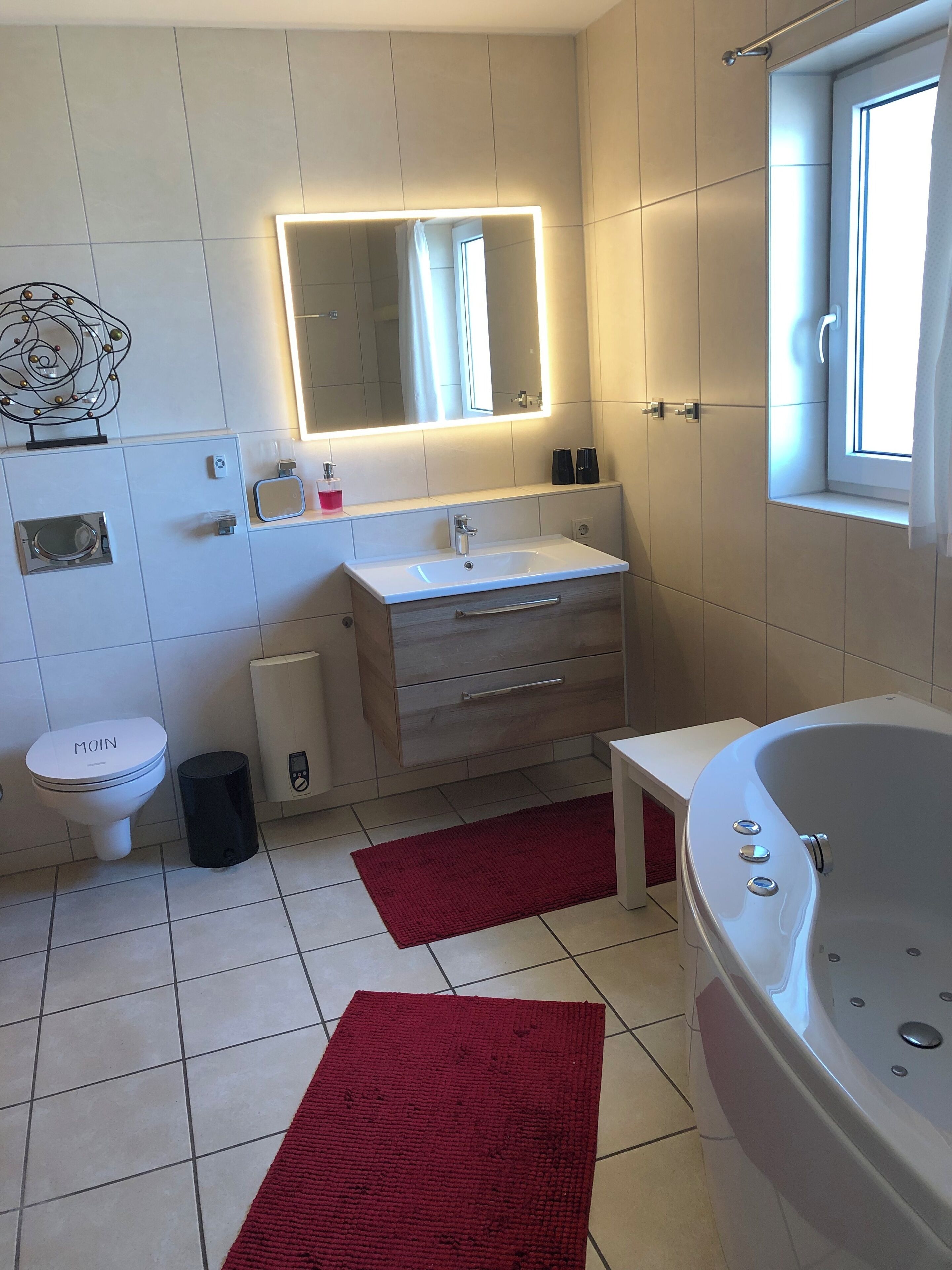 Een moderne badkamer met een witte wastafel, een badkamermeubel en een groot bad. Er is een toilet, een prullenbak en een rode vloerkleed.