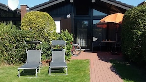 A building with a dark roof and a large glass door. There are two lounge chairs in the front yard. - Vacation home bungalow, one floor, fenced garden, dog allowed, 2 parking spaces (Butjadingen)