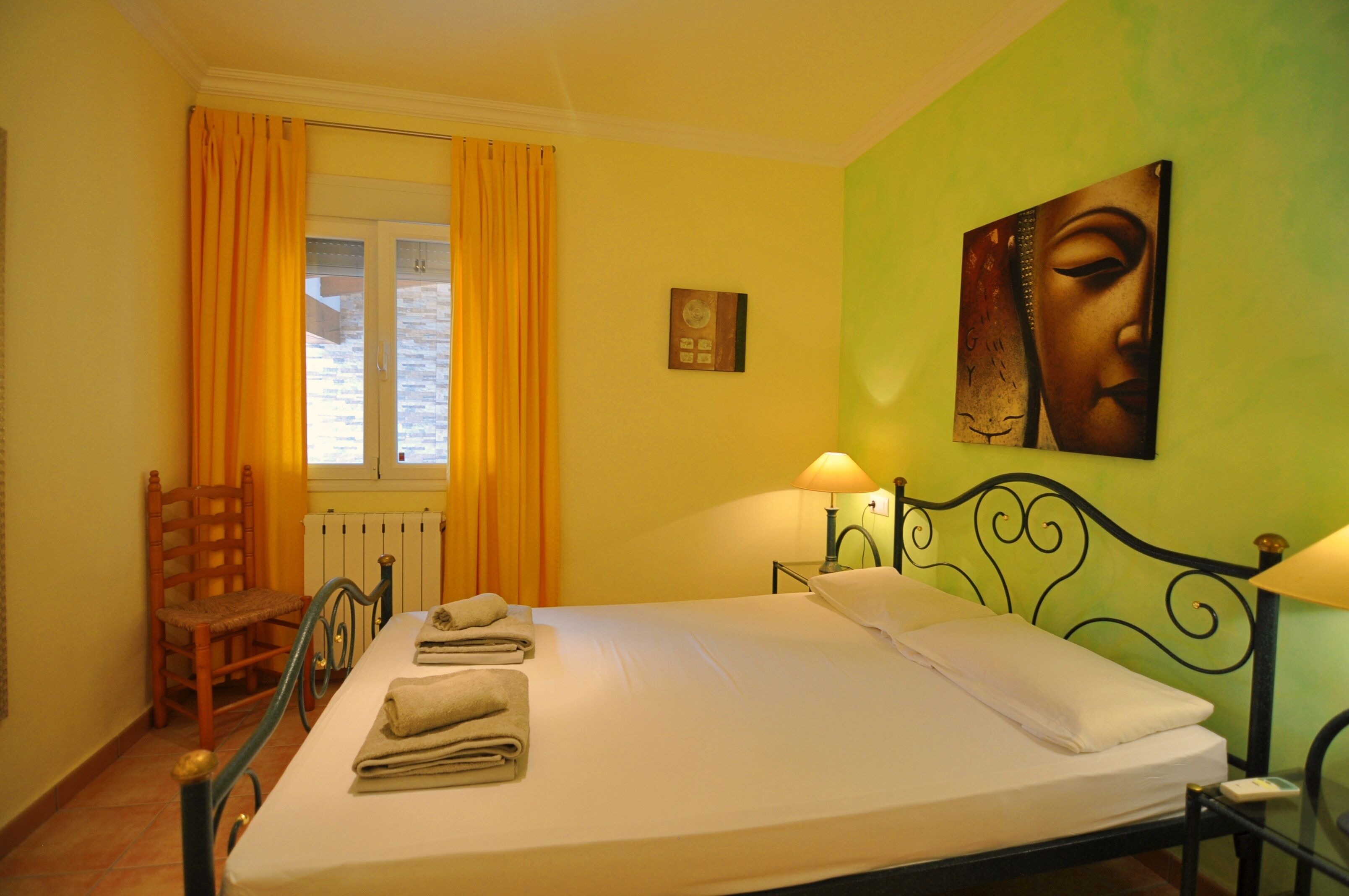 A hotel room with a bed, a chair, a window with curtains, a wall painting, and a lamp.