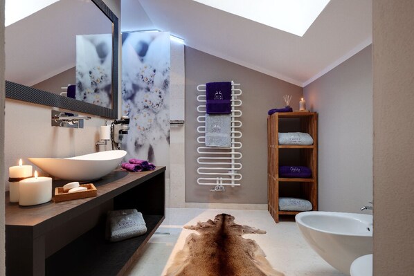 A modern bathroom with a large skylight, a freestanding bathtub, a vanity with a mirror, and a wooden shelf with towels.