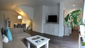 Un salon moderne avec un canapé, une table basse et un téléviseur. Un escalier mène à un autre niveau, et il y a un coin repas avec une table et des chaises.