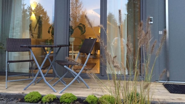 Un bâtiment moderne doté de grandes portes vitrées, d’un espace détente avec deux fauteuils et une table, et d’un petit jardin avec des plantes.