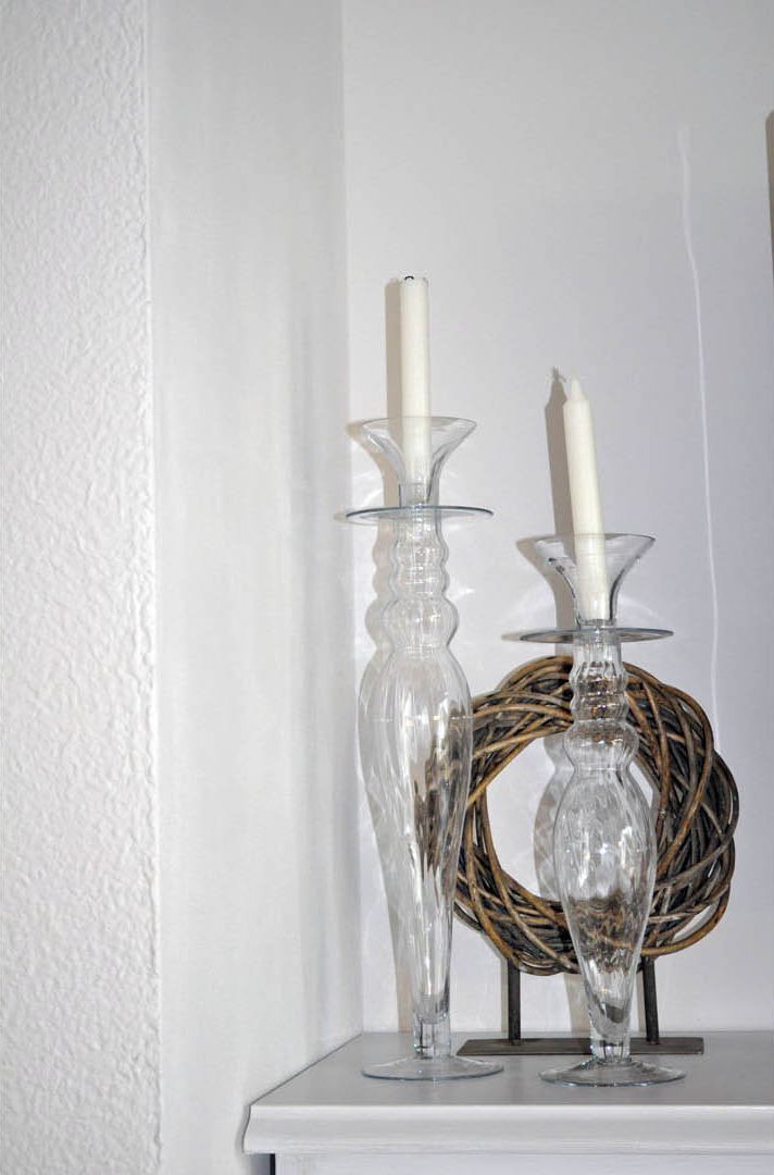 Two clear glass candle holders with white candles on a white shelf against a textured white wall.