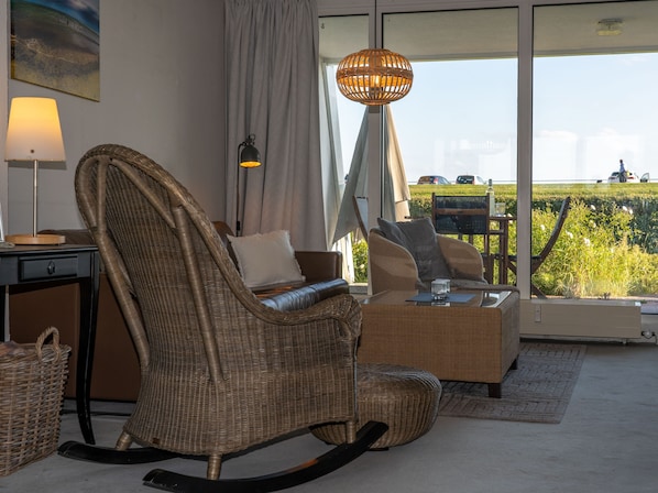 A room with wicker furniture, a lamp, a glass table, and a view of a green landscape through large windows. - 096 Havenkante - 096 Hafenkante apartment with harbor view and direct beach access (Wilhelmshaven)