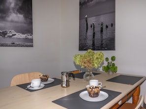 Ein Esstisch mit einer weißen Tasse, einem Metallkrug, einer Vase mit grünen Blumen und einem Muffin auf einem Untertaler.