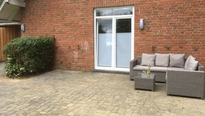 Un bâtiment en briques doté d’une grande porte-fenêtre coulissante, d’un patio avec un canapé en osier et une table, et d’un petit arbuste sur la gauche.