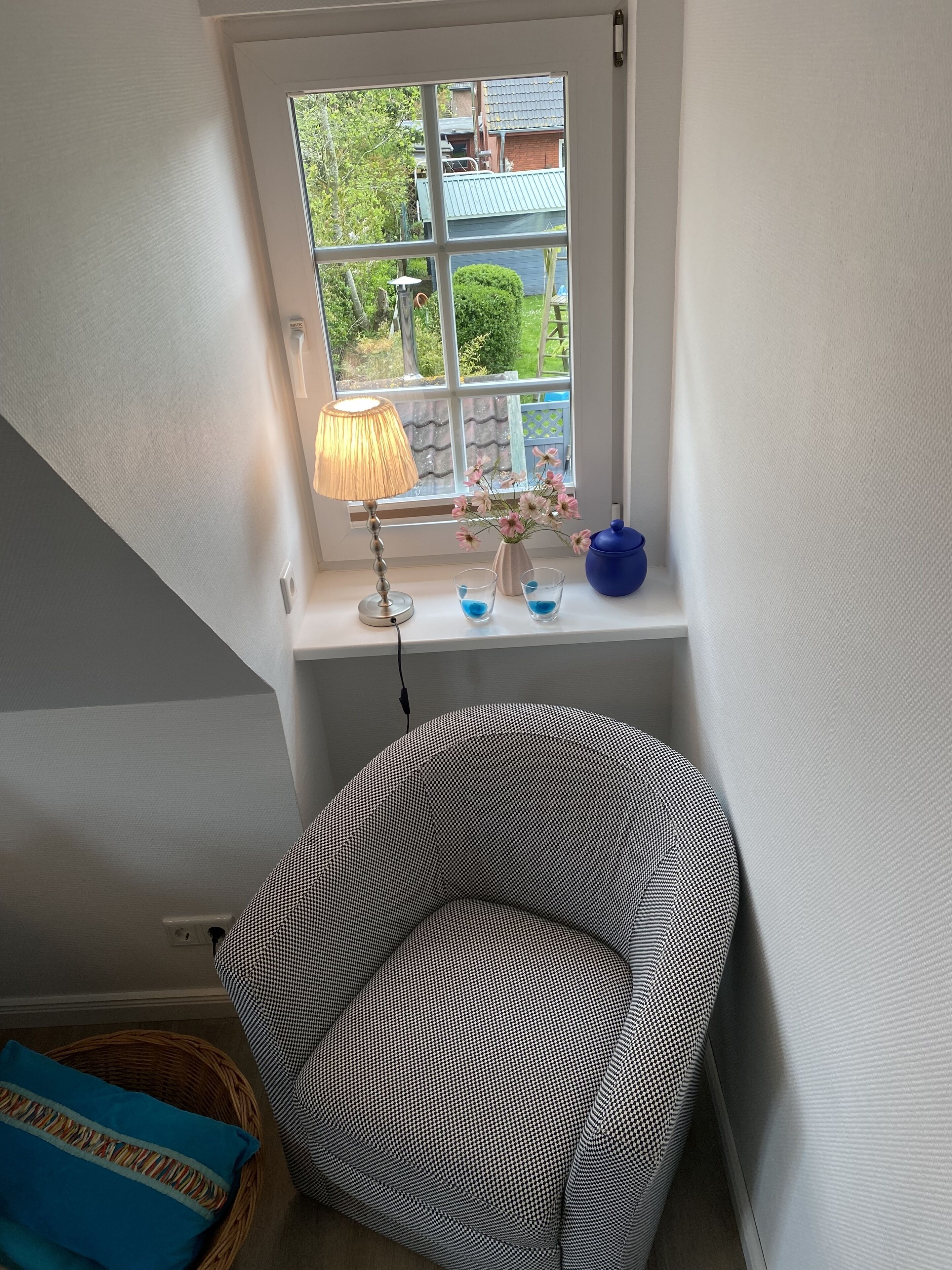 Una habitación de esquina con ventana, una lámpara, una mesita con un jarrón de flores y una silla estampada.