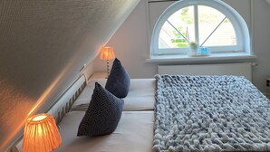Quarto com teto inclinado, cama com edredom branco, duas almofadas estampadas, manta de tricô, abajur e janela com vista para vegetação.