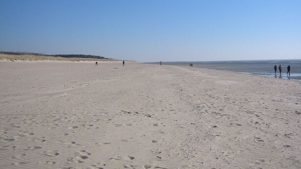 Ampla praia de areia com pegadas e algumas pessoas caminhando.