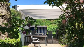 A covered patio area with a table and chairs, surrounded by lush greenery and a tree. - FH / Central to the Baltic Sea/North Sea/Denmark/Sylt/Amrum/Föhr (Stadum)