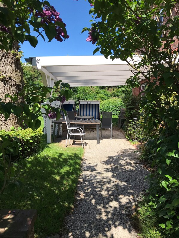 A covered patio area with a table and chairs, surrounded by lush greenery and a tree.