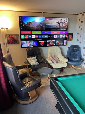 A living room with a pool table, two leather chairs, a small round table, and a flat-screen TV mounted on the wall displaying various channels.