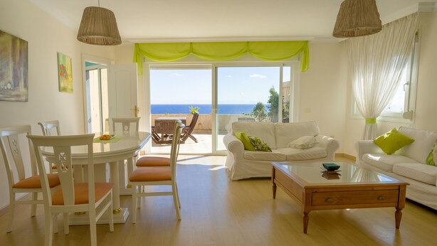 Un salón espacioso con comedor, un sofá blanco con cojines verdes, una mesa de centro de madera y vistas al mar a través de grandes ventanales.
