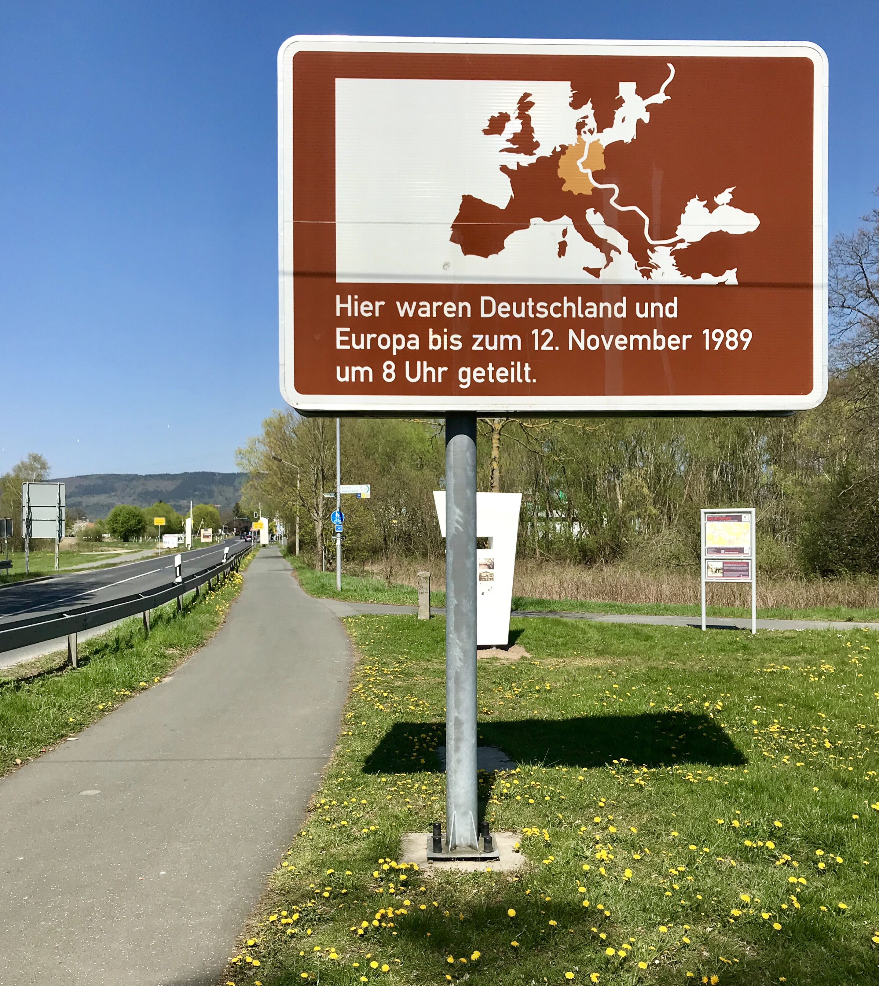 Ein Wegweiser mit einer Karte und Text in einer Fremdsprache, neben einer Straße mit Gehweg und Grünflächen.