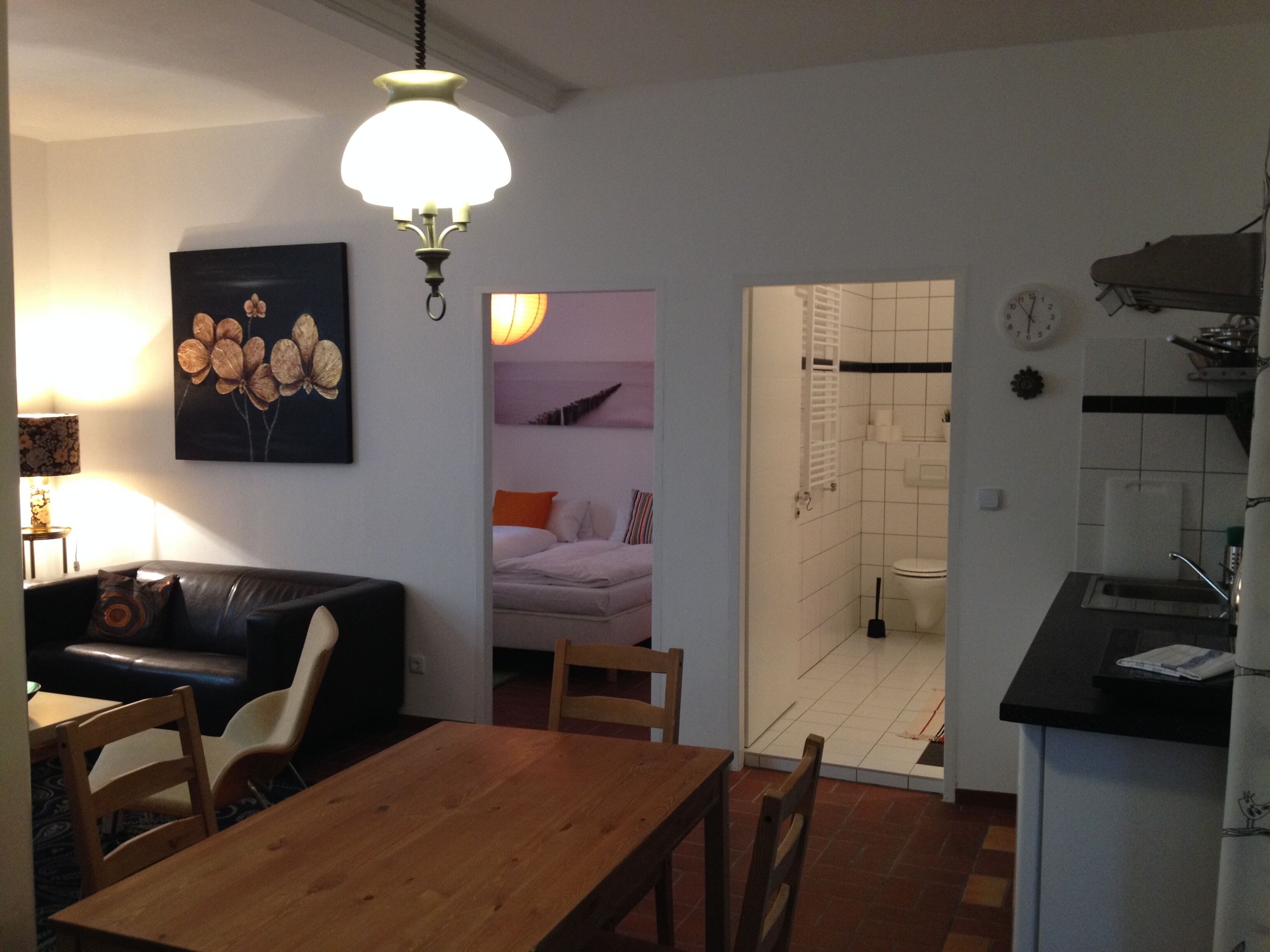 Ein Wohnzimmer mit Holztisch und -stühlen, einer schwarzen Ledersofa sowie einer Küchenzeile mit Spüle und Herd. An der Wand hängt ein Gemälde.
