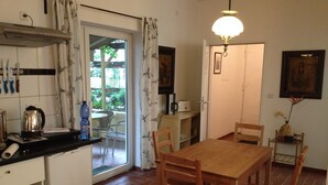 Une cuisine avec un coin repas, une fenêtre avec des rideaux, une table, des chaises, une bouilloire et une bouteille d’eau.