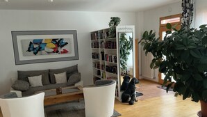 Un salon moderne avec un canapé gris, des fauteuils blancs, une bibliothèque et une grande plante en pot.