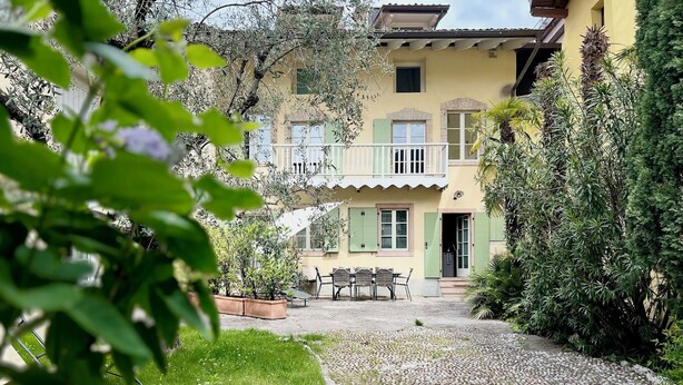 A two-story building with a balcony, a patio with a table and chairs, and a gravel pathway. - Lake Garda: Idyllic villa in quiet village center with olive and palm trees garden (Nago-torbole)