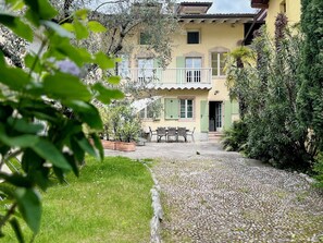 Un bâtiment à deux étages doté d’un balcon, d’une terrasse avec une table et des chaises, et d’un chemin de gravier.